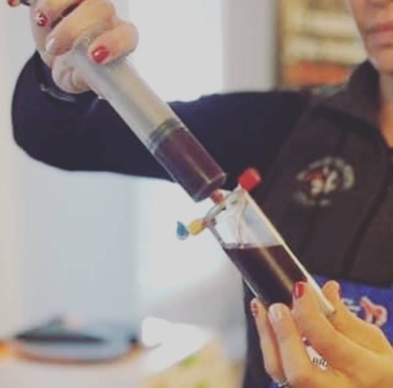 A person with red-painted nails is holding a tube and transferring a dark red liquid, resembling blood, from a syringe into a small vial with two colored stoppers at an animal hospital.