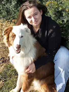 A woman with long brown hair, wearing a black sweater and light blue jeans, sits outdoors on grass, hugging a large, fluffy brown and white dog in the sunlight—perhaps after a happy visit to the veterinarian. Shrubs are in the background.