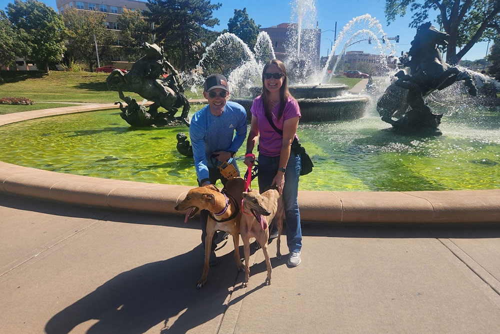 A man and woman smiling and posing with a brown greyhound dog in front of a large, ornate fountain with statues and water sprays on a sunny day.
