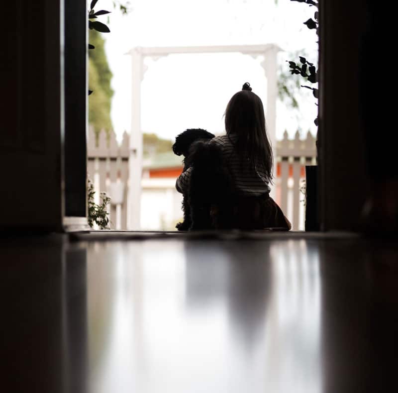 A young girl sits at an open doorway, hugging a small black dog as they gaze out toward a bright picket fence and garden—capturing a quiet moment that could be seen in any caring veterinarian’s home. The softly lit floor gleams in the foreground.